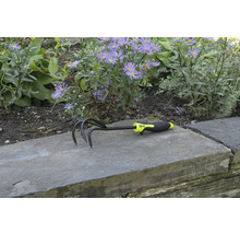 Cultivator de grădină cu trei dinți și mâner din plastic în fața unui strat de flori