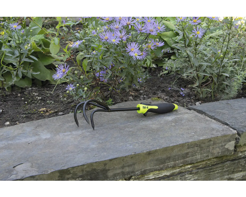 Cultivator de grădină cu trei dinți și mâner din plastic în fața unui strat de flori