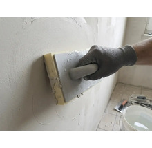 Mână cu mănușă de lucru folosind o drișcă cu burete pentru a netezi tencuiala pe un perete.