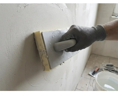 Mână cu mănușă de lucru folosind o drișcă cu burete pentru a netezi tencuiala pe un perete.