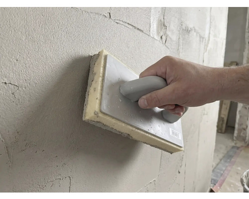 Mână care folosește un drisc cu burete de 28 pe 14 centimetri pentru a netezi tencuiala proaspătă pe un perete.
