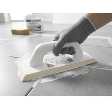 O persoană cu mănușă de protecție aplică chit de rosturi alb pe plăci de gresie gri folosind o dristă pentru rășină epoxidică.