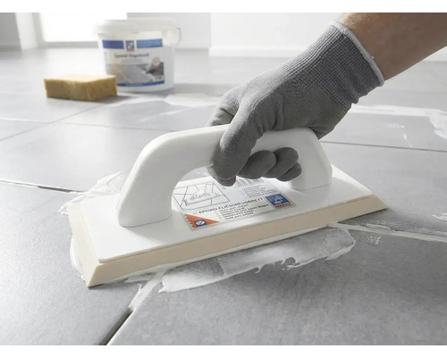 O persoană cu mănușă de protecție aplică chit de rosturi alb pe plăci de gresie gri folosind o dristă pentru rășină epoxidică.