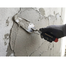 O mână cu mănușă de lucru aplică mortar pe un perete cu o mistrie Maurerlob.