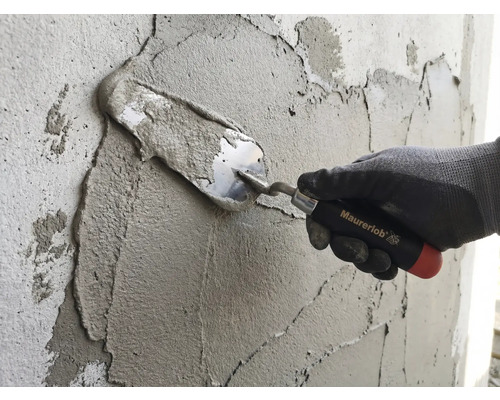 O mână cu mănușă de lucru aplică mortar pe un perete cu o mistrie Maurerlob.