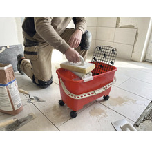 O persoană care stă în genunchi curăță plăcile cu o drișcă cu burete și un set de spălare roșu pe roți.