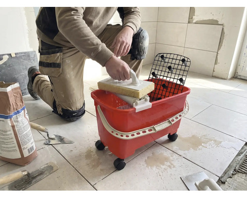 O persoană care stă în genunchi curăță plăcile cu o drișcă cu burete și un set de spălare roșu pe roți.