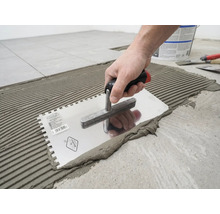 O mână aplică adeziv pentru plăci ceramice pe podea cu o gletieră cu dinți, creând un model cu caneluri.