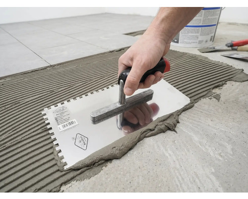 O mână aplică adeziv pentru plăci ceramice pe podea cu o gletieră cu dinți, creând un model cu caneluri.