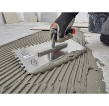 Mână cu mănușă de lucru aplică adeziv pentru gresie pe pardoseală cu o gletieră cu dinți din oțel inoxidabil.