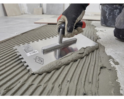 Mână cu mănușă de lucru aplică adeziv pentru gresie pe pardoseală cu o gletieră cu dinți din oțel inoxidabil.