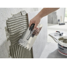 O mână aplică adeziv pentru plăci ceramice pe un perete cu o gletieră cu dinți.