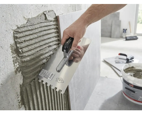 O mână aplică adeziv pentru plăci ceramice pe un perete cu o gletieră cu dinți.