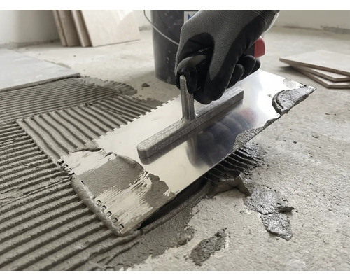 O persoană aplică adeziv pentru gresie pe o podea de beton folosind o mistrie dințată din metal.