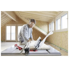 Un bărbat cu ochelari de protecție taie o scândură de lemn cu un ferăstrău circular cu masă într-o cameră luminoasă cu grinzi de lemn.