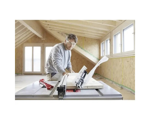 Un bărbat cu ochelari de protecție taie o scândură de lemn cu un ferăstrău circular cu masă într-o cameră luminoasă cu grinzi de lemn.