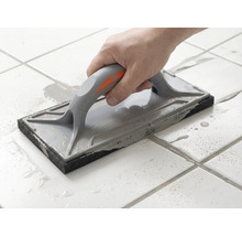 O mână ghidează o drișcă pentru rosturi peste plăci de gresie albe pentru a întinde chitul de rosturi gri.