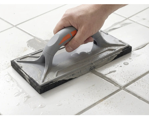 O mână ghidează o drișcă pentru rosturi peste plăci de gresie albe pentru a întinde chitul de rosturi gri.