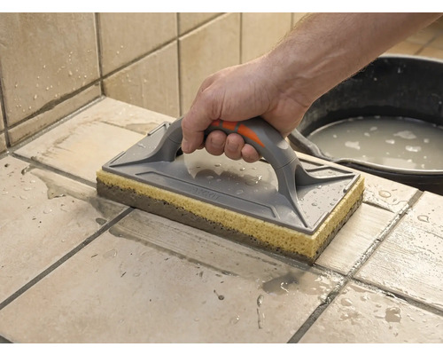 Drișcă cu buret folosită pentru curățarea plăcilor de faianță proaspăt chituite. O mână ghidează drișca pe plăcile ude.