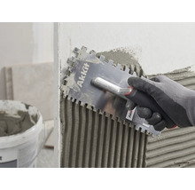 O mână cu mănușă aplică uniform adeziv pentru plăci pe un perete cu o gletieră cu dinți Akkit.