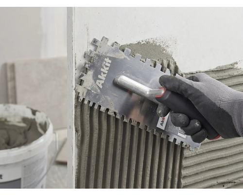 O mână cu mănușă aplică uniform adeziv pentru plăci pe un perete cu o gletieră cu dinți Akkit.