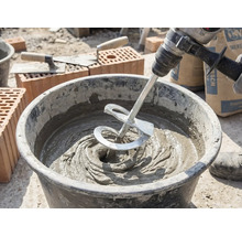 Un amestecător pe o bormașină amestecă mortar gri într-o găleată pe un șantier cu cărămizi și mistrii.