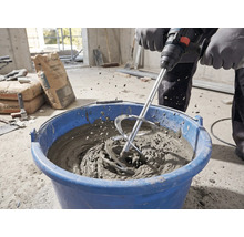 O persoană amestecă mortar cu un agitator și un malaxor într-o găleată albastră pe un șantier.