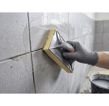 O mână curăță plăcile de faianță proaspăt chituite cu o drișcă cu burete și apă.