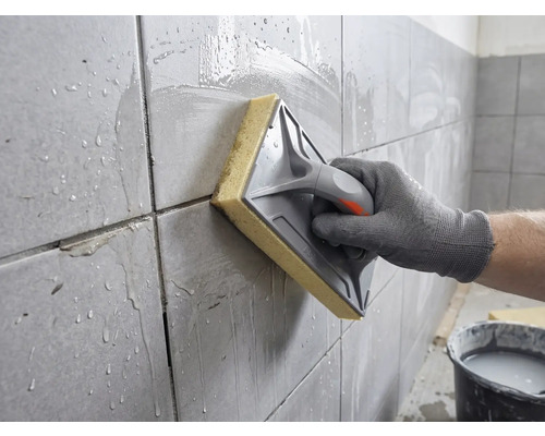 O mână curăță plăcile de faianță proaspăt chituite cu o drișcă cu burete și apă.