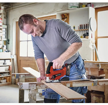 Un meșter taie o scândură de lemn pe un banc de lucru într-un atelier luminos folosind un ferăstrău pendular cu acumulator Einhell. Einhell Logo.