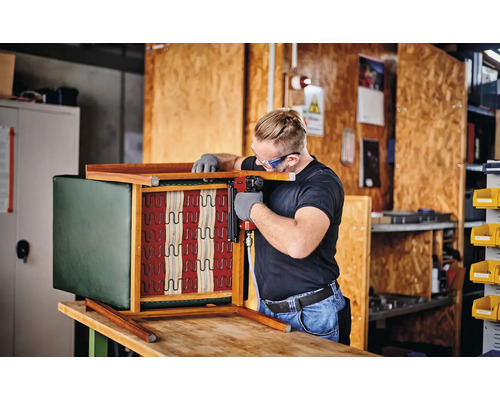 Meșter care fixează tapițeria unui scaun de lemn cu un capsator pneumatic într-un atelier.