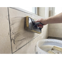 O persoană curăță plăci de faianță bej cu o drișcă cu burete. O găleată albă cu apă se află în prim-plan.