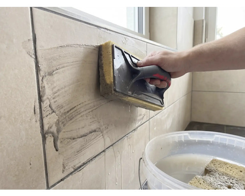 O persoană curăță plăci de faianță bej cu o drișcă cu burete. O găleată albă cu apă se află în prim-plan.