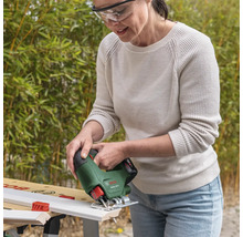 Femeie cu ochelari de protecție taie o șipcă albă cu un ferăstrău vertical Bosch EasySaw 18 Volt pe un banc de lucru într-o grădină luminoasă.