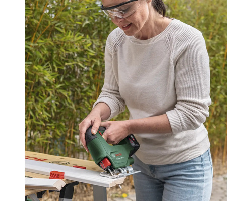 Femeie cu ochelari de protecție taie o șipcă albă cu un ferăstrău vertical Bosch EasySaw 18 Volt pe un banc de lucru într-o grădină luminoasă.