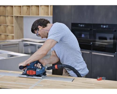 Bărbat care utilizează un ferăstrău circular cu șină de ghidare și aspirare a prafului pe un blat de lemn într-o bucătărie.