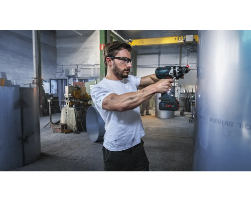 Meșter cu ochelari de protecție care utilizează o mașină de găurit cu acumulator Bosch ProCore 18 Volt pe o țeavă de metal într-un atelier industrial.