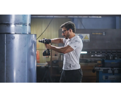 Un bărbat găurește o țeavă mare de metal cu o mașină de găurit cu acumulator într-un atelier. Poartă ochelari de protecție.