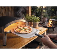 O pizza proaspăt coaptă este scoasă dintr-un cuptor portabil pe o masă de lemn în grădină cu o paletă pentru pizza cu Tenneker Logo.