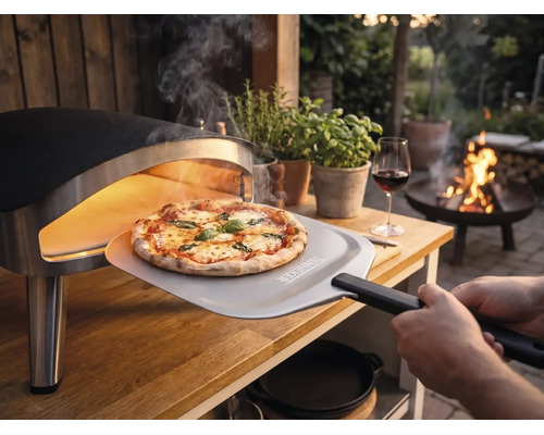 O pizza proaspăt coaptă este scoasă dintr-un cuptor portabil pe o masă de lemn în grădină cu o paletă pentru pizza cu Tenneker Logo.