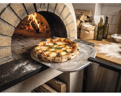 Pizza proaspăt coaptă pe o paletă pentru pizza cu Tenneker Logo în fața unui cuptor de piatră aprins într-o bucătărie rustică.