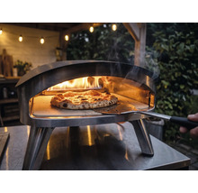 Cuptor de pizza din oțel inoxidabil pentru exterior cu flăcări și o pizza pe piatră de pizza, manevrată cu o paletă pentru pizza.