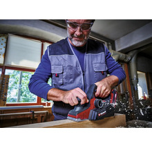 Un bărbat cu ochelari de protecție folosește o rindea cu acumulator cu Einhell Logo pe o grindă de lemn într-un atelier luminos și organizat.