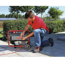 Un bărbat îngenunchează pe o terasă și conectează un cablu la un generator de curent mobil Einhell.