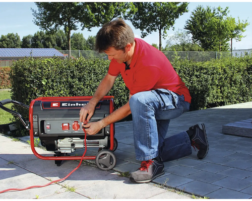 Un bărbat îngenunchează pe o terasă și conectează un cablu la un generator de curent mobil Einhell.