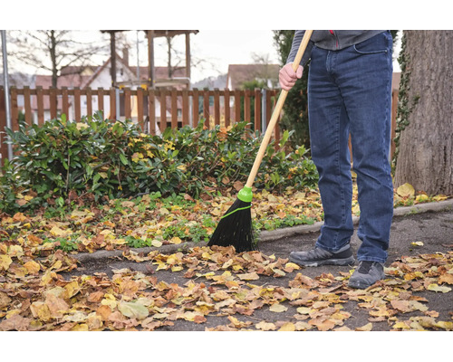 O persoană folosește o mătură de grădină cu coadă de lemn și peri din plastic pentru a curăța frunzele de toamnă de pe o alee asfaltată.