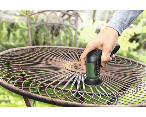 O mână folosește un șlefuitor multifuncțional Bosch cu acumulator pentru a șlefui un scaun de grădină rotund din metal în exterior.