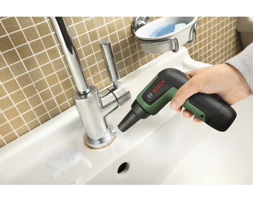 O mână utilizează o perie de curățare cu acumulator Bosch pentru a curăța baza unei baterii de chiuvetă cromate.