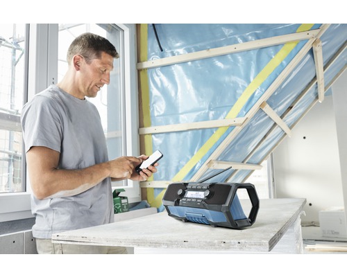 Un bărbat operează un telefon mobil lângă un radio de șantier într-o cameră cu pereți neterminați