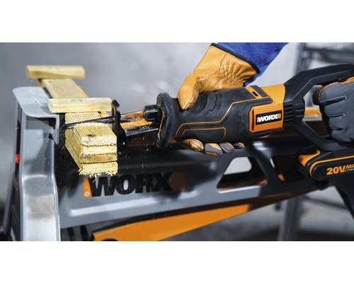 Fierăstrău electric cu acumulator tăind lemn pe o masă de lucru, operat de o persoană cu mănuși, logo-ul Worx vizibil.